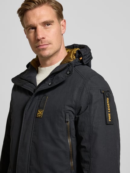 Männermodel trägt Parka mit Label-Detail van PME Legend in zwart met gele details, zichtbaar ritssluiting, Nahaufnahme Oberkörper, Regular Fit, capuchon, borstzakken met ritssluiting, patzakken, context: ruglengte 89 cm maat M, schouderbreedte 50 cm, borstwijdte 58 cm, mouwlengte 86 cm.