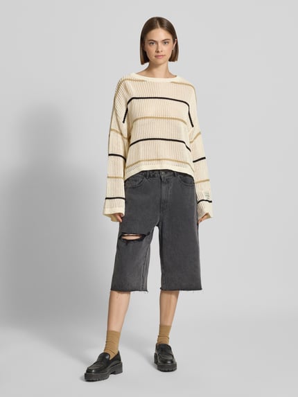 Frauenmodel trägt Oversized Strickpullover mit Label-Detail und Rundhalsausschnitt von Karo Kauer, Grobstrick, Streifenmuster in Creme und Schwarz, Kombi mit dunkler Shorts, Socken und Loafern, Ganzkörperansicht von vorne.