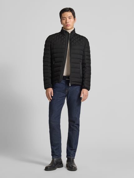 Męski model nosi Slim Fit Steppjacke mit gefüttertem Kragen von Christian Berg Men, czarny kolor, krój slim fit, kieszenie zapinane na zamek błyskawiczny, podszewka na kołnierzu, elastyczne zakończenia rękawów, długość tyłu w rozmiarze M: 68 cm, szerokość ramion w rozmiarze M: 47 cm, szerokość klatki piersiowej w rozmiarze M: 52 cm, długość rękawów w rozmiarze M: 68 cm, widok całej sylwetki z przodu
