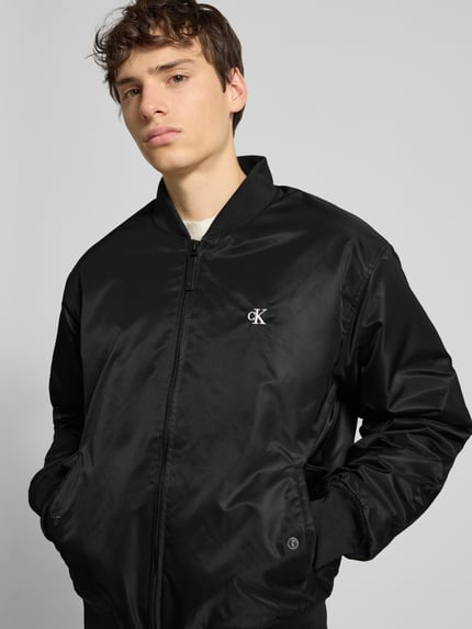 Männermodel trägt Bomberjacke mit Label-Stitching von Calvin Klein Jeans in Schwarz, Regular-Fit, Stehkragen, Reißverschluss, Eingrifftaschen, Nahaufnahme Oberkörper. Rückenlänge 68 cm, Schulterbreite 60 cm, Brustweite 62 cm, Ärmellänge 68 cm.