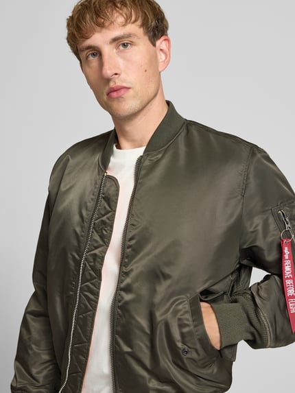 Männermodel trägt Bomberjacke mit Pattentaschen und Label-Applikation von Alpha Industries in Olivgrün, glatte Textur, Nahaufnahme Oberkörper. Regular Fit, Stehkragen, Reißverschluss, Maße: Rückenlänge 69 cm, Schulterbreite 44 cm, Brustweite 54 cm, Ärmellänge 64 cm.