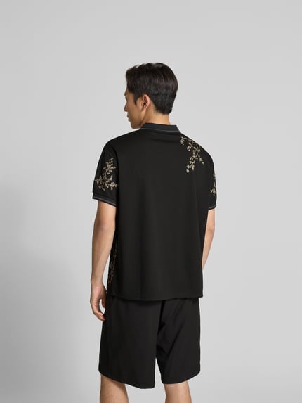 Męski model nosi Poloshirt floralem Allover-Stitching od Don´t Waste Culture, czarny kolor, florystyczny haft, krój relaxed fit, logo z nadrukiem, kołnierz polo, krótki rękaw, długość tyłu w rozmiarze M: 71 cm, zdjęcie całej sylwetki z tyłu.