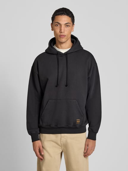 Männermodel trägt Relaxed Fit Hoodie aus Baumwoll-Mix Modell 'JET BL' von Levi's® in Schwarz, beige Hose, Ganzkörperansicht von vorne.