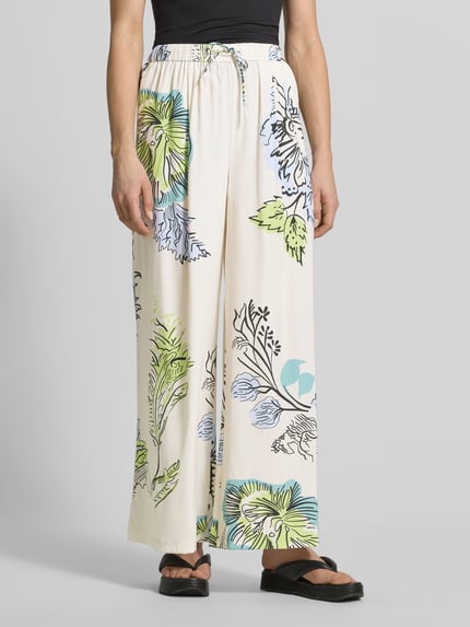 Vrouwenmodel draagt Stoffhose aus Seide-Mix van HERZENS, lichte stof, loose fit, elastische tailleband met tunnelkoord, bloemenprint, vooraanzicht, context: binnen beenlengte maat 36 is 76 cm, taillebreedte maat 36 is 37 cm.