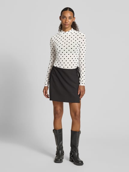 Frauenmodel trägt Slim Fit Langarmshirt mit Allover-Muster Modell 'JESHI' von PIECES, weiß mit schwarzen Punkten, Ganzkörperansicht von vorne.