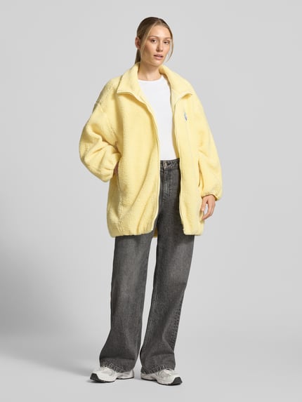 Frauenmodel trägt Oversized Jacke mit Stehkragen Modell 'Hoktown' von American Vintage in Gelb, Fleece, Reißverschluss, Logo-Patch, Ganzkörperansicht von vorne.