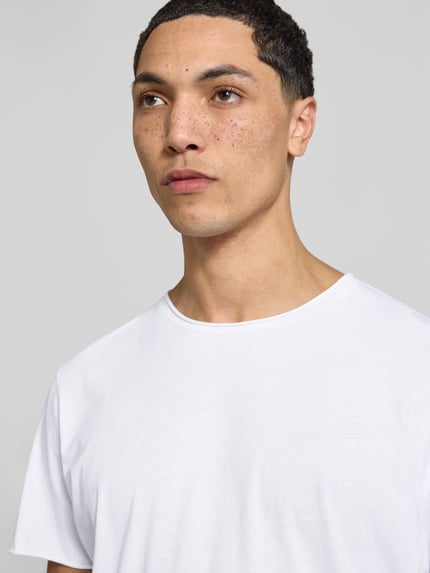 Męski model nosi T-Shirt mit Rollsaum od Filippa K z czystej bawełny, prosty krój, okrągły dekolt, zrolowany brzeg, długość tyłu w rozmiarze M: 70 cm, zbliżenie na górną część ciała.