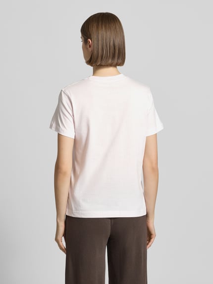 Modelka nosi T-Shirt mit Label-Stitching und Rundhalsausschnitt von New Balance, bawełna, krój regular fit, okrągły dekolt, długość tyłu w rozmiarze S: 56 cm, jasne kolory, prosty krój, gładka faktura, całkowity widok od tyłu.
