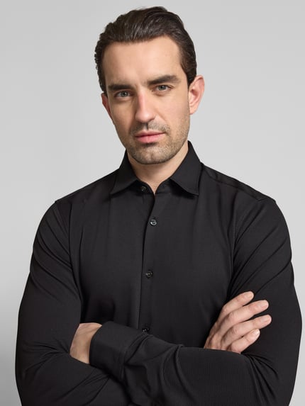 Mężczyzna prezentuje Slim Fit Businesshemd in Struktur-Optik Modell 'ERMO' von HUGO, kolor czarny, pika, kołnierzyk typu kent, wyhaftowane logo, listwa guzikowa na całej długości, długi rękaw, sportowy mankiet z regulacją. Nahaufnahme Oberkörper.
