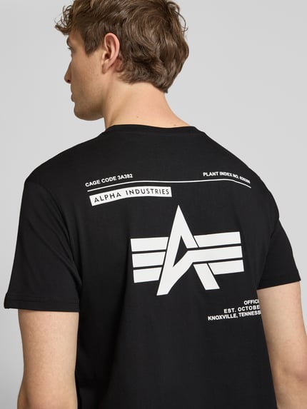 Männermodel trägt T-Shirt mit Label-Patch von Alpha Industries in Schwarz, große weiße Aufdrucke auf glatter Baumwolle, Rückenansicht, Reine Baumwolle, Gerader-Schnitt, Rundhalsausschnitt, Herren T-Shirt, Rückenlänge bei Größe M: 66 cm. Ganzkörperansicht Rückansicht.