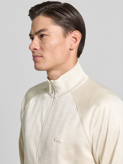 Männermodel trägt Regular Fit Sweatjacke mit Logo-Patch von Lacoste in Beige mit feiner Textur und Reißverschluss, Nahaufnahme Oberkörper. Herren Sweatjacke von Lacoste