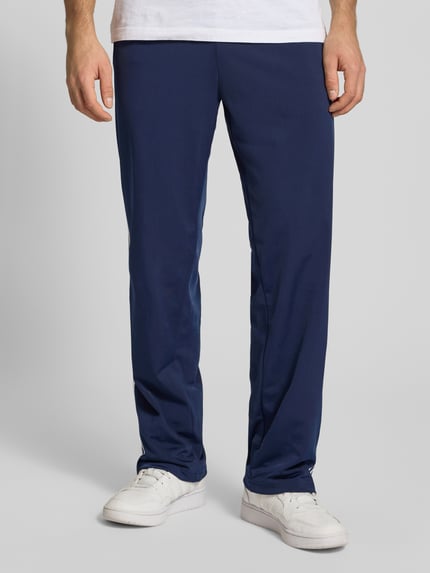 Męski model nosi Trainingshose mit Kontraststreifen und Reißverschlusstaschen od adidas Originals, krój regular fit, elastyczny pas, model FIREBIRD, paski w kontrastowym kolorze, wyhaftowane logo, kieszenie zapinane na zamek błyskawiczny, długość wewnętrzna nogawki 78 cm, rozmiar M, granatowe, gładka tekstura, całkowity widok z przodu.