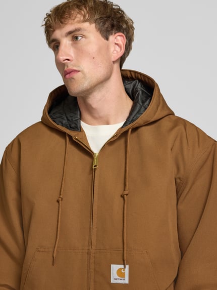 Männermodel trägt Jacke mit Label-Patch und Känguru-Tasche von Carhartt Work In Progress in Braun, goldener Reißverschluss, Nahaufnahme Oberkörper. Reine Baumwolle, Kapuze mit Kordelzug, Regular Fit, gerippte Abschlüsse, Bildkontext integriert.