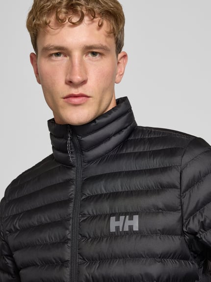 Männermodel trägt Steppjacke mit Label-Print von Helly Hansen, schwarze Farbe, Stehkragen, Nahaufnahme Oberkörper.