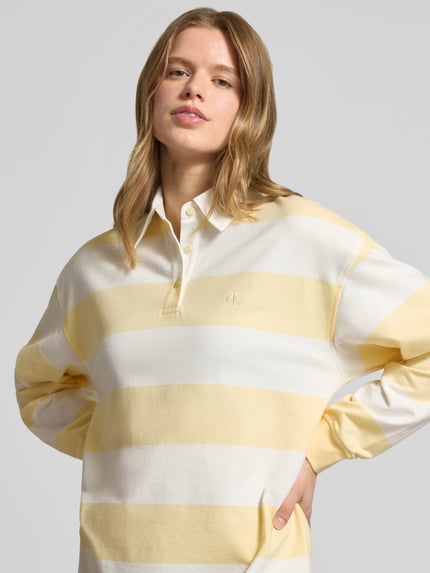 Modelka kobieca nosi Sukienka mini z kołnierzykiem polo model 'Varsity' od Calvin Klein Jeans, krój regular fit, bawełna, wyhaftowane logo, elastyczne zakończenia rękawów, paski biało-żółte, szerokość w rozmiarze S: 52 cm, długość tyłu: 84 cm, długość rękawów: 53 cm, zbliżenie na górną część ciała.