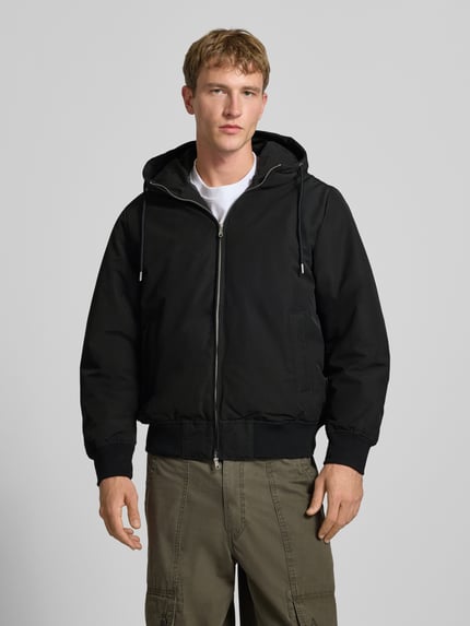 Męski model nosi czarną Jacke mit Kapuze und Eingrifftaschen Modell 'TRACE' von JACK & JONES, krój regular fit, widok całej sylwetki z przodu. Szerokość w rozmiarze M: 60 cm, długość tyłu: 70 cm, szerokość ramion: 46 cm, długość rękawa: 68 cm.