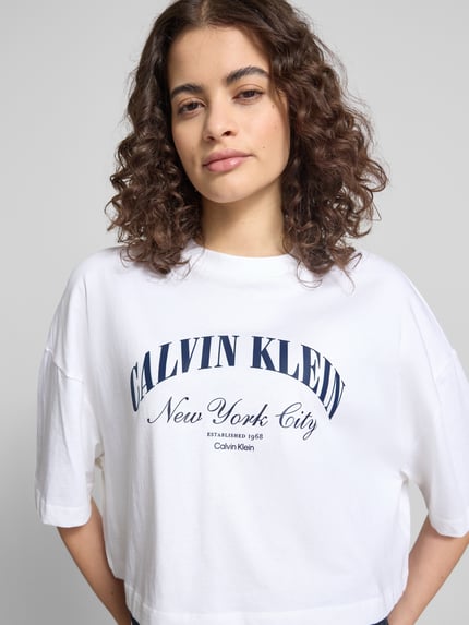 Kobieta modelka nosi Cropped T-Shirt mit Logo-Print od Calvin Klein Jeans z bawełny, krój krótki, nadruk z logo, okrągły dekolt, długość tyłu w rozmiarze S: 42 cm, zdjęcie: zbliżenie na górną część ciała