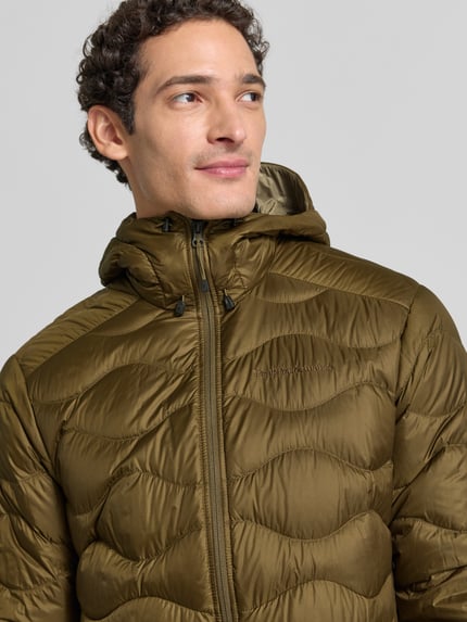 Mężczyznamodel nosi Regular Fit Steppjacke mit Kapuze Modell 'Helium Down Hood' von Peak Performance, oliwkowy kolor, wyhaftowane logo, krój regular fit, kaptur z tunelem, zamek błyskawiczny, Szerokość ramion w rozmiarze M: 42 cm, Szerokość klatki piersiowej w rozmiarze M: 55 cm, Długość tyłu w rozmiarze M: 71 cm, Długość rękawów w rozmiarze M: 70 cm, ujęcie: zbliżenie na górną część ciała