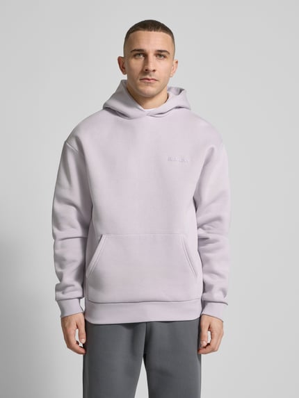 Męski model nosi Hoodie mit Känguru-Tasche von REVIEW w kolorze jasnoszarym, mieszanka bawełny, luźny krój, kaptur, detal z logo, kieszeń kangurka, elastyczne wykończenia, długość tyłu w rozmiarze M: 68 cm, długość rękawów w rozmiarze M: 62 cm, całkowity widok z przodu.