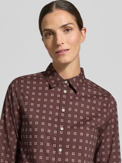 Frauenmodel trägt Longline Langarmbluse mit Label-Detail von GANT, braun mit geometrischem Allover-Muster, Nahaufnahme Oberkörper.