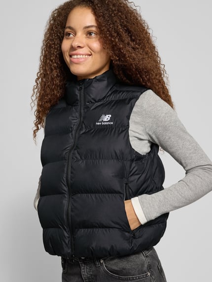 Frauenmodel trägt Steppweste mit Stehkragen Modell 'BOSTON PUFFER VEST' von NEW BALANCE in Schwarz mit Logo-Print, Struktur, Reißverschlusstaschen, Ganzkörperansicht von vorne