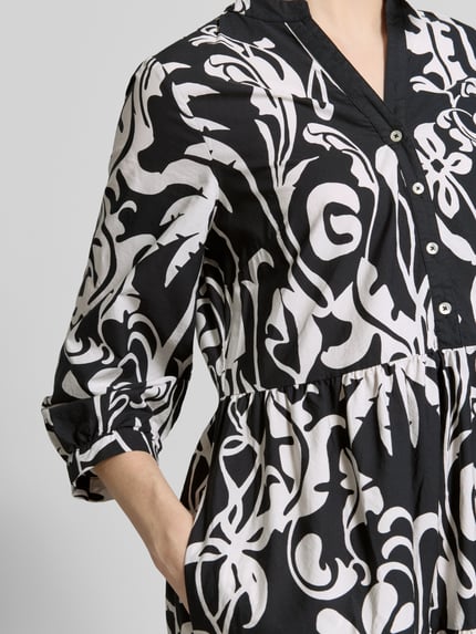 Vrouwenmodel draagt 'Midikleid mit Eingrifftaschen und 3/4-Arm' van Emily Van den Bergh, zwart-witte allover motief, katoen, V-hals, knoopsluiting, A-lijn, steekzakken, 3/4-mouw, detailopname bovenlichaam.