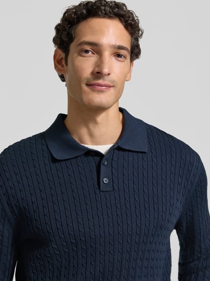 Mężczyzna nosi sweter z dzianiny 'Strickpullover mit Polokragen und kurzer Knopfleiste' od MCNEAL, kolor granatowy, ścieg warkoczowy, kołnierzyk polo, listwa guzikowa, bawełna, krój regular fit, prążkowane wykończenia, długość tyłu 71 cm, długość rękawów 78 cm, ujęcie: zbliżenie górnej części ciała.