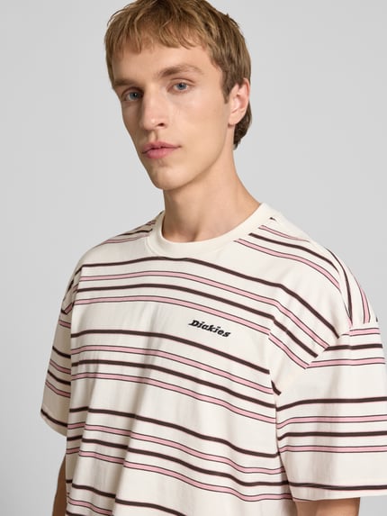 Męski model nosi T-Shirt z logo-stitching model 'EAST LIBERTY' od Dickies, bawełna, krój relaxed fit, kremowe i bordowe paski, zbliżenie górnej części ciała.