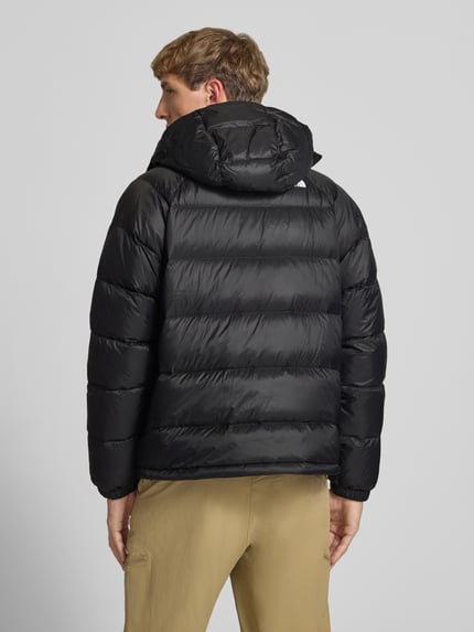 Männermodel trägt Jacke mit Logo-Stitching und Kapuze von The North Face in Schwarz, gesteppt, gerader Schnitt, Ganzkörperansicht Rückansicht. Herren Jacke, Kapuze, Reißverschluss, seitliche Reißverschlusstaschen, Rückenlänge 66 cm, Schulterbreite 45 cm, Brustweite 55 cm, Ärmellänge 70 cm.