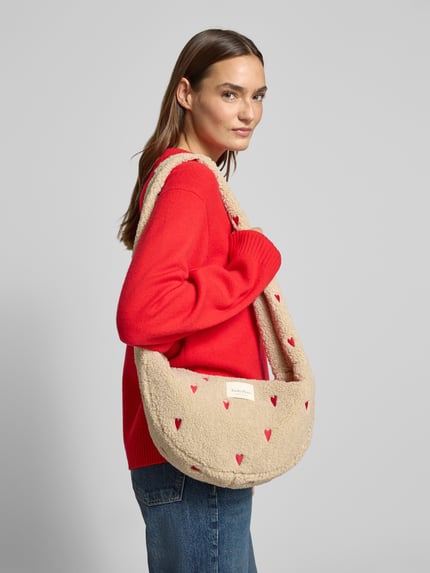 Frauenmodel trägt Shoulder Bag mit Teddyfell Modell 'Hearts' von STUDIO NOOS in Beige mit roten Herzmotiven, detailreiche Struktur, Nahaufnahme Oberkörper. Hauptfach mit Reißverschluss, Logo-Patch, Innentasche, Maße 42x21x13 cm, Schulterriemen 43 cm.
