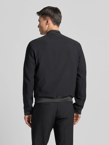 Mężczyzna model nosi Blouson mit seitlichen Eingrifftaschen od MCNEAL, czarny kolor, elastyczne wykończenia, materiał z domieszką stretchu, krój regular fit, stójka, zamek, szerokość w rozmiarze M: 60 cm, szerokość ramion w rozmiarze M: 45 cm, długość tyłu w rozmiarze M: 65 cm, długość rękawa w rozmiarze M: 71 cm, tylna pełna sylwetka