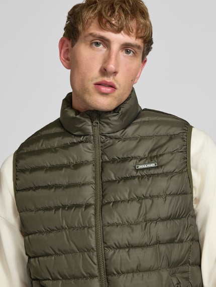 Mężczyzna nosi kamizelkę pikowaną z zamkiem błyskawicznym Model 'ELVIS' od JACK & JONES, oliwkowy, detal z logo, krój regular fit, stójka, kieszenie zapinane na zamek błyskawiczny, długość tyłu w rozmiarze M: 67 cm, szerokość ramion w rozmiarze M: 41 cm, szerokość klatki piersiowej w rozmiarze M: 54 cm, zbliżenie na górną część ciała.