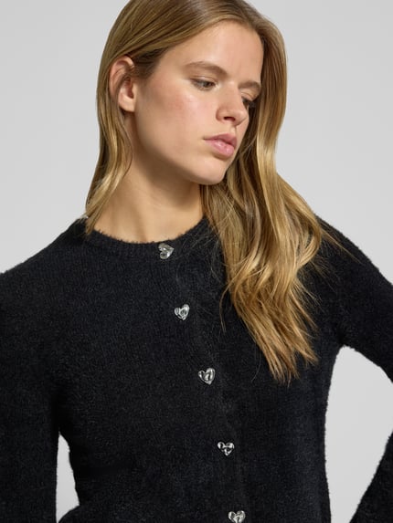 Frauenmodel trägt Strickjacke mit Herzknöpfen von ONLY, schwarzer Stoff, silberfarbene Knöpfe, detailreiche Textur, Nahaufnahme Oberkörper.