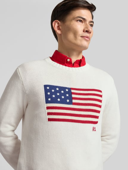 Männermodel trägt Strickpullover mit Rundhalsausschnitt von Polo Ralph Lauren in Weiß mit US-Flaggenmotiv aus Baumwolle, Nahaufnahme Oberkörper.