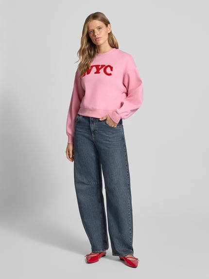 Frauenmodel trägt Relaxed Fit Sweatshirt aus Baumwoll-Mix Modell 'Tatum' von JJXX in Rosa mit rotem Statement-Patch, Damen Sweatshirt mit Rippbündchen, überschnittenen Schultern, langärmligem Design und Rundhalsausschnitt, weiche Textur und lockerer Schnitt, Ganzkörperansicht von vorne.