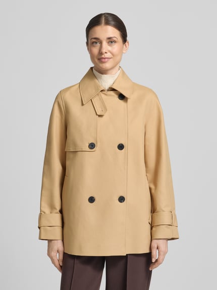 Frauenmodel trägt Trenchcoat mit Ärmelriegeln von Stefanel in Beige mit schwarzen Knöpfen, Umlegekragen, Zweireiher, Eingrifftaschen, Reine Baumwolle, Regular Fit, Ganzkörperansicht von vorne.