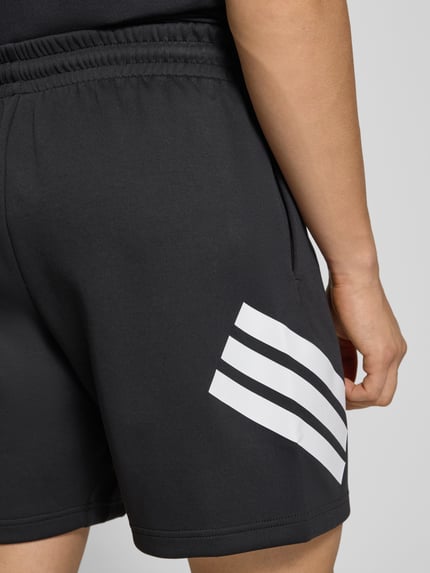Männermodel trägt Regular Fit Sport-Shorts mit Logo-Print von adidas Sportswear in Schwarz, seitliche Eingrifftaschen, Nahaufnahme Oberschenkel.