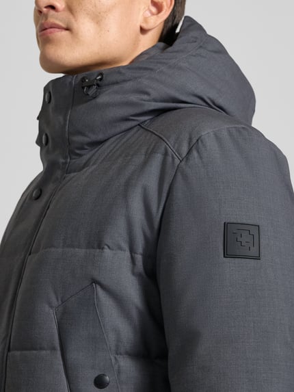 Männermodel trägt Daunenjacke mit Kapuze von Strellson in unifarbener Optik, sichtbarer Logo-Patch, Struktur, Nahaufnahme Oberkörper.