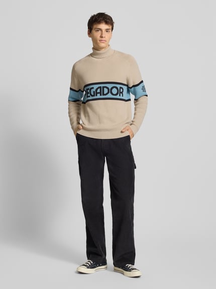 Mannenmodel draagt Strickpullover met Rollkragen van Pegador in beige met blauwe accenten, katoenmix, geribde randen, labelprint, regular fit, vooraanzicht volledige lichaamsopname.