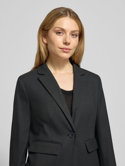 Modelka kobieca nosi Slim Fit Blazer mit Reverskragen od More & More, czarny, materiał z domieszką wiskozy, zapięcie na guzik, kieszenie z patkami, widok na górną część ciała z przodu.