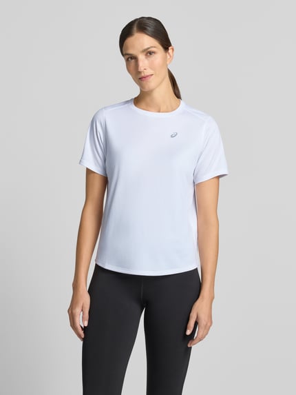 Frauenmodel trägt T-Shirt mit Rundhalsausschnitt und Logo-Print von ASICS in Weiß, gerader Schnitt, leicht abgerundeter Saum, Ganzkörperansicht von vorne.