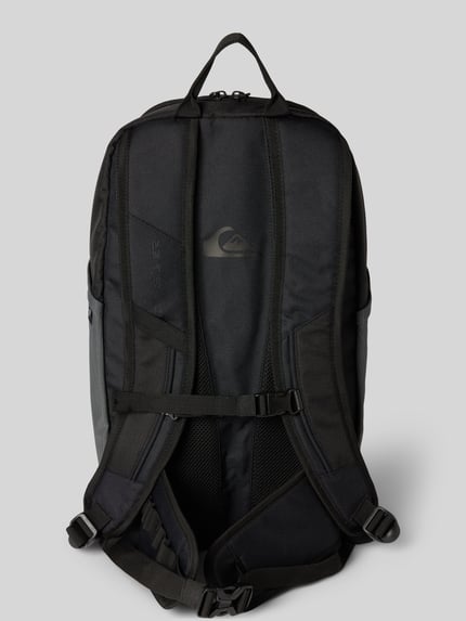 Männermodel trägt Rucksack mit Logo-Patch Modell 'ALTACAMA' von Quiksilver in Schwarz mit verstellbaren Schultergurten und Tragegriff, Ganzkörperansicht Rückansicht.