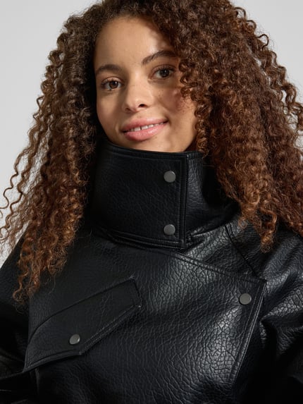 Kobiece modelka nosi czarną kurtkę w stylu skóry Jacke in Leder-Optik Modell 'SILLE' od Topshop, krój regular fit, wysoki kołnierz, patki na kieszeniach, pasek, ozdobne napy, tekstura skóropodobna, Szerokość w rozmiarze S: 56 cm, Długość pleców: 80 cm, Szerokość ramion: 44 cm, Długość rękawa: 73 cm, zbliżenie na górną część ciała.