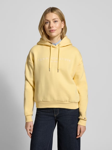 Vrouwenmodel draagt Relaxed Fit Hoodie aus Baumwoll-Mix van Tommy Hilfiger in effen geel, katoenmix sweatstof, label stitching, langemouw, ribboorden, Ganzkörperansicht von vorne.