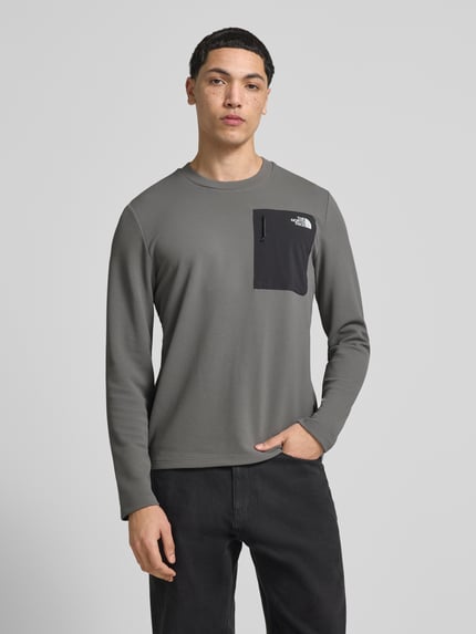 Männermodel trägt Langarmshirt mit Napoleontasche Modell 'KECHA' von The North Face, Elastischer-Material-Mix, Gerader-Schnitt, Rundhalsausschnitt, Label-Detail, Rückenlänge 67 cm bei Größe M, graue und schwarze Farbakzente, Ganzkörperansicht von vorne.