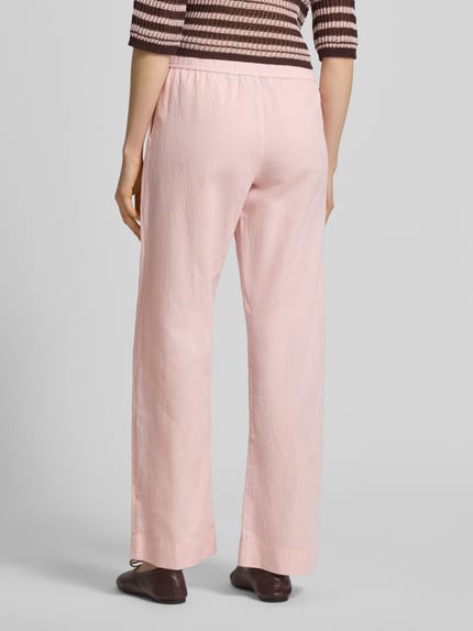 Frauenmodel trägt Stoffhose mit Leinen-Anteil und Eingrifftaschen von Neo Noir, ausgestellter Schnitt, Modell 'Sonar', Lockerer-Schnitt, pastellrosa, feine Textur, Ganzkörperansicht Rückansicht. Damen Stoffhose aus Baumwoll-Mix mit Leinen-Baumwoll-Anteil, elastischer Bund, französische Eingrifftaschen, Bundweite 36 cm, Innenbeinlänge 79 cm, Modell Neo Noir.