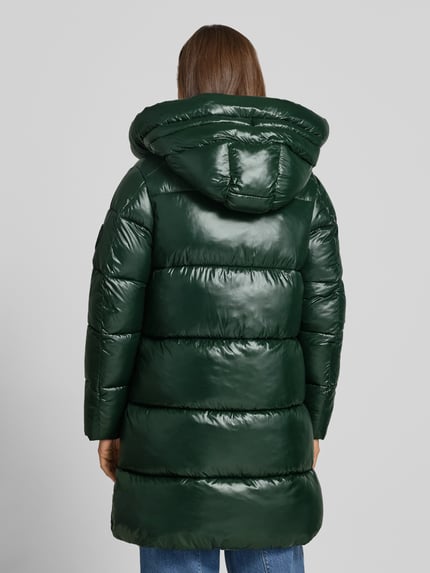 Frauenmodel trägt Steppmantel mit Kapuze und Label-Patch Modell 'ISABEL' von Save The Duck, glänzend grün, Ganzkörperansicht Rückansicht. Gerade geschnitten mit Tunnelzugkapuze, Zweiwege-Reißverschluss und seitlichen Reißverschlusstaschen.