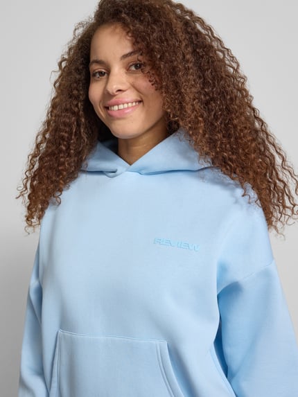 Frauenmodel trägt Hoodie mit Logo-Print und Känguru-Tasche von REVIEW in Hellblau, Baumwoll-Mix, Loose-Fit, Kapuze, Nahaufnahme Oberkörper.