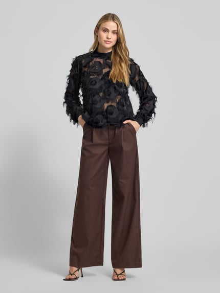 Vrouwenmodel draagt semitransparante zwarte blouse met franjes, opstaande kraag, knoopsluiting achteraan, Regular Fit, model 'DIO' van YAS. Volledige lichaamsweergave van voren.