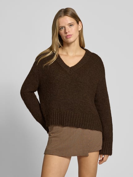 Frauenmodel trägt Pullover mit tiefem V-Ausschnitt von ONLY in Dunkelbraun, strukturierte Wolloptik, kombiniert mit kariertem Rock, Ganzkörperansicht von vorne. Damen Pullover von ONLY